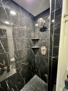 Luxurious black marble-look walk-in shower with rainfall showerhead, handheld sprayer, built-in corner shelves, and dark hexagon floor tiles, completed as part of custom bathroom remodeling in Utah.