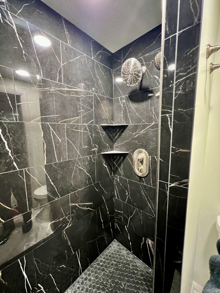 Luxurious black marble-look walk-in shower with rainfall showerhead, handheld sprayer, built-in corner shelves, and dark hexagon floor tiles, completed as part of custom bathroom remodeling in Utah.